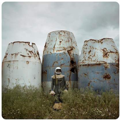 A person wearing a helmet and dark suit with straw fringes stands in tall grass in front of three large, rusted cylindrical tanks, under a stormy sky