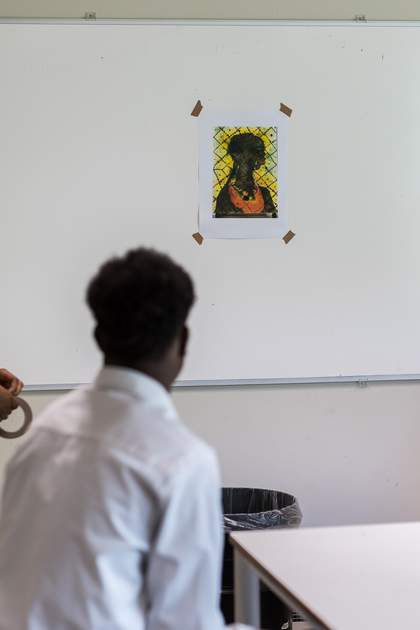 A teenage boy faces the whiteboard, which has a printed out copy of Chris Ofili’s ‘No Woman, No Cry’ taped to it. His back is to the camera. He wears a white shirt.