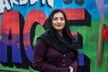 A portrait of teacher Mia Bano. She is standing outside in front of a colourful mural. She wears a dark purple cardigan and a black headscarf.
