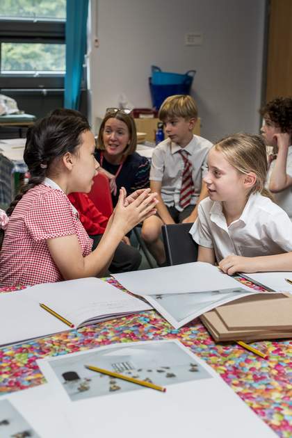 Two young people sit at a table with their notebooks open. They face each other and are having a discussion. A printout of the artwork sits on the table between them.