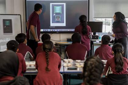 Students look at Parviz Tanavoli’s ‘Cage, Cage, Cage’ projected in their classroom on a digital screen. Two students stand at the front of the class, facing the artwork.