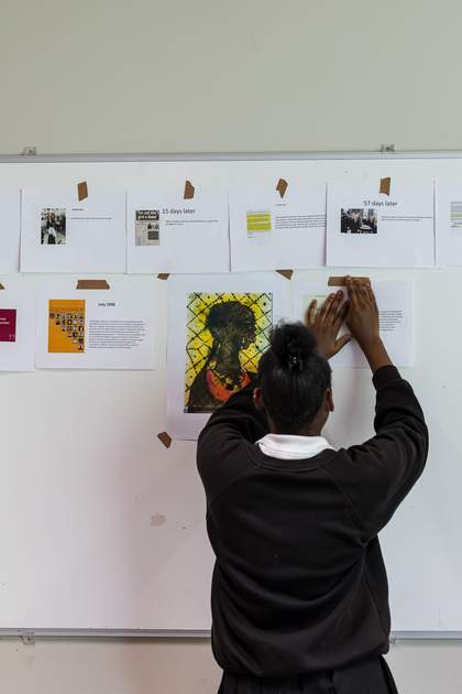 A student tapes a piece of paper with text on it to the whiteboard. Together with the other images and pieces of text, it forms a timeline of the Stephen Lawrence case.