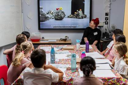 Students sit around a table, watching Tate’s film about Edgar Calel. The film shows fruits, vegetables and flowers sitting on rocks in a gallery space. Teacher Lyndsey Miles sits to the right of the screen, looking up at it as well.