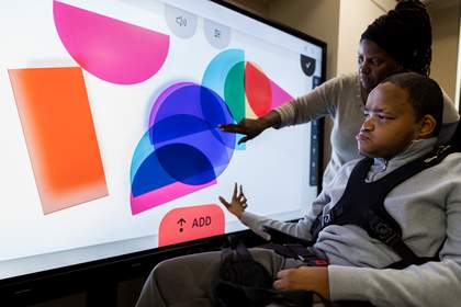 A student is interacting with large coloured shapes on a large interactive whiteboard in a classroom.