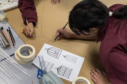 A student leans over a piece of cardboard, using a pencil to sketch the outline of a birdcage. Another student draws the hook at the top of the cage. They look at a reference picture of four birdcages. To their left are rolls of tape, straws, and a ruler.