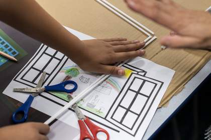 A student places straws overtop of their drawing of a scene outside a window. The scene has a rainbow, a large tree, and some people playing. Two pairs of scissors sit on top of the sheet of paper.
