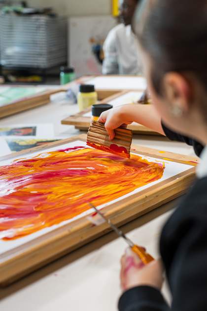 A student uses a strip of corrugated cardboard to paint swirls of red, orange and yellow onto a canvas. They hold scissors in their left hand.