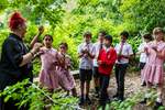 Teacher Lyndsey Miles stands with students in a forest clearing. The students are holding clay sculptures in their hands. The student nearest Lyndsey has their hand raised to ask a question.
