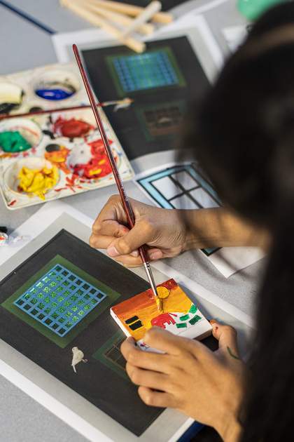 A young person paints a tiny canvas. There is a red elephant, a red and yellow sky, and some green areas for the ground. Paint pots and printouts of Parviz Tanavoli's 'Cage, Cage, Cage' are visible on the table behind them.