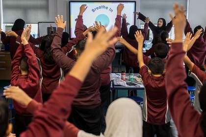 Mia stands in front of the class, raising her arms up in the air as part of their ‘Rainbow Breathing’ mindfulness exercise. Students face her, mimicking her arm movement. The digital screen at the front of the classroom reads, ‘Breathe In.’