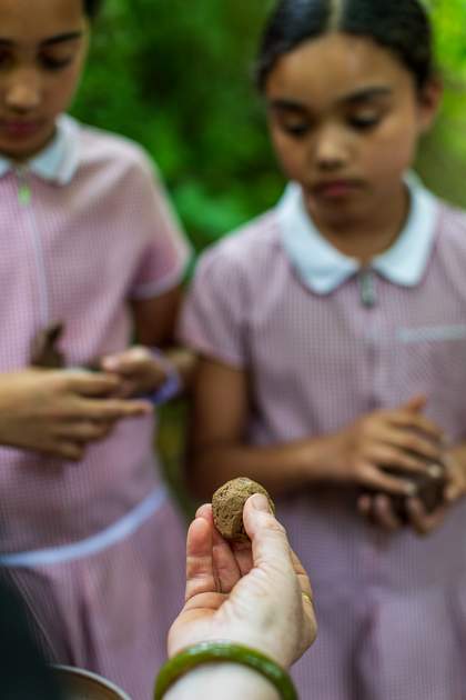Lyndsey holds a seed ball in her fingers. Two students in the background look down at it as they hold their clay vessels.