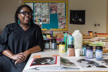 Nakessa Simpson sits at a table in her art classroom. She is looking off to her left and smiling. She wears a black shirt, long turquoise earrings and thick framed black glasses. On the table are bottles of paint and photocopied portraits of Black figures