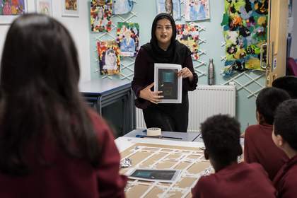 Mia stands in front of students, holding a printed out picture of Parviz Tanavoli’s ‘Cage, Cage, Cage.’ In front of her, on the table, is a birdcage structure made out of tape and straws.