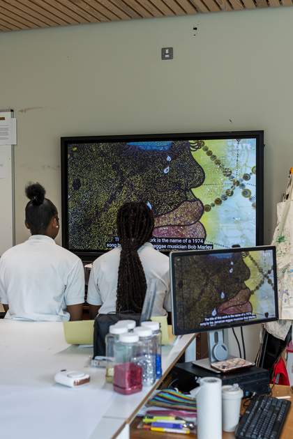 Two teenage girls watch the Tate Shots film about ‘No Woman, No Cry’ on their video screen. The video shows a close-up of the work and the caption mentions the work's title is from a song by Bob Marley.