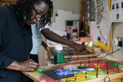 A teacher bends over an artwork she's making. It has a painted background of red, green and yellow, covered with diagonal black lines that create a diamond pattern. A photocopied face has been pasted on. A box of beads sits on top of the photocopy.