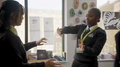 Two students face each other in an art classroom, mirroring each others hand movements and gestures. Student artwork is displayed on the walls and windows behind them