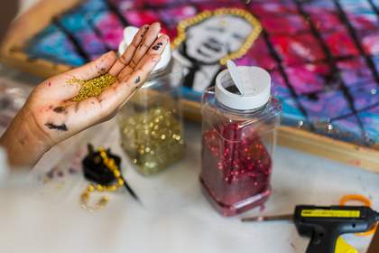 A student’s left hand, covered in paint, is filled with gold glitter. Jars of gold and red glitter, as well as gold beads, a glue gun, and an artwork are visible in the background.