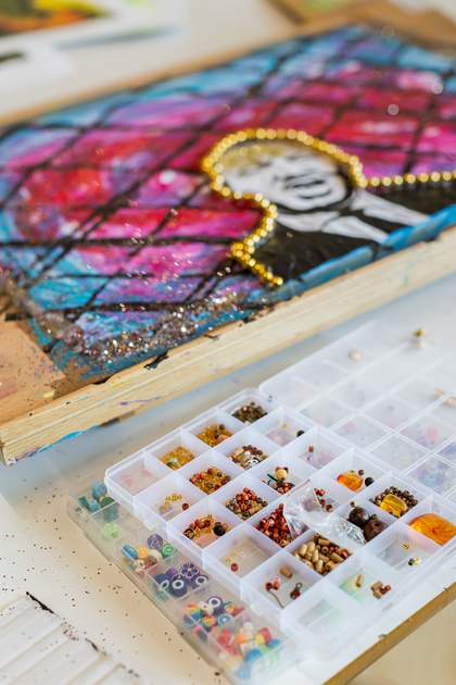 An artwork and a box of beads sit on a table. The artwork has a painted background with diagonal black lines that create a diamond pattern. A black and white picture of Martin Luther King, Jr. has been pasted in the centre, outlined with gold beads.