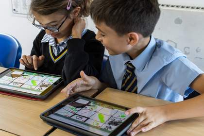 Two students sat at desks in a classroom are exploring a collaborative drawing game on tablet devices.