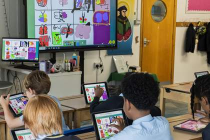A group of students sat at desks in a classroom are making colourful drawings on tablet devices with the drawing also displayed on a large screen at the front of the room