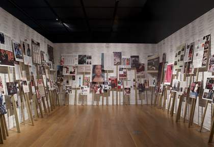 A room with white walls and wooden floor. The three walls of the room visible have placards of different sizes and heights visible. The placards have images of women from magazines on them.