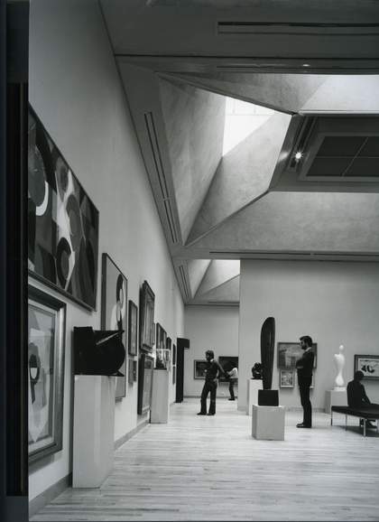 Black and white image of a gallery with sculptures and paintings in a modern looking gallery. There are a couple of people looking at the artworks.