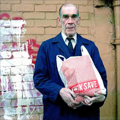 A photograph of a man holding a red and white striped Kwik Save bag. He is wearing a blue jacket with a dark tie and shirt.
