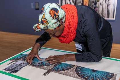 A teacher leans over a large sheet of paper, sticking down photocopies of colourful patterns to make a collage