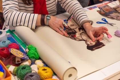 A teacher sticks down a printed photograph of a woman onto a large roll of paper while holding scissors and colourful sellotape. Around the paper are many colourful balls of string and other art materials, ready to be used in the activity.