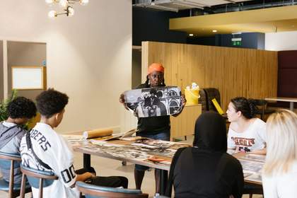 A photograph a woman holding a black and white image whilst talking to students sat at a table.