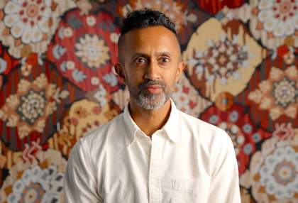 a man with brown skin, wearing a white shirt sits in front of a colourful background