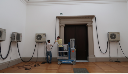 Two individuals holding up a long wire that connects several wall-mounted fans together and runs along the top of a large doorway flanked by pillars