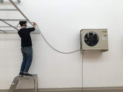An individual standing on a small platform and holding up a long wire that connects to a fan mounted on the wall