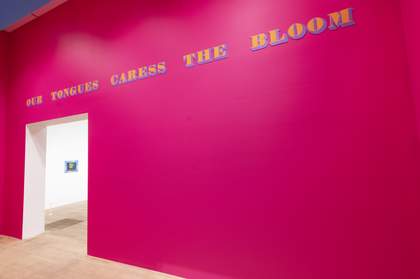 A bright pink wall with the words ‘Our tongues caress the bloom’ written on it in large letters, above an archway that leads into a white gallery space