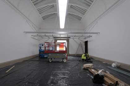 A view of an art gallery with a metal grid installed horizontally across it above head height, and black plastic sheeting, cardboard boxes and industrial trolleys underneath it