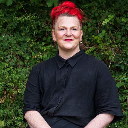 A portrait of teacher Lyndsey Miles. Behind her is a forest filled with bright green leaves, which contrasts with her bright red hair.