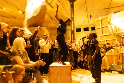 Photograph of nora chipaumire singing into a microphone, whilst dancer Yinka Esi Graves stands atop a wooden box. They are surrounded by a large audience watching.
