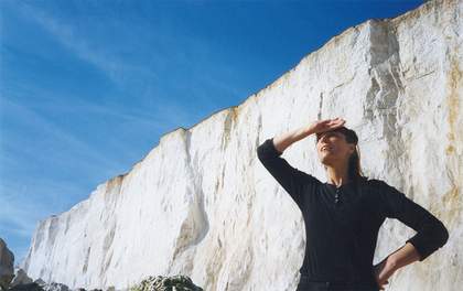 Cornelia Parker stands in front of the White Cliffs of Dover on a sunny day. She holds her right hand up to shield her face from the sun. Her left hand is on her hip.