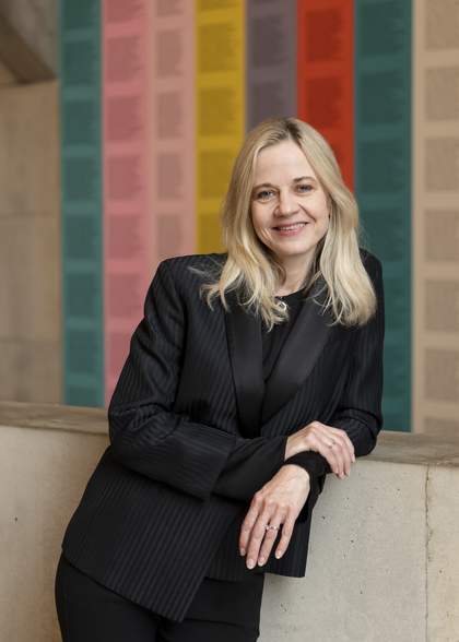 photo of a light skinned woman with light hair in a black jacket standing in front of a wall with vertical stripes