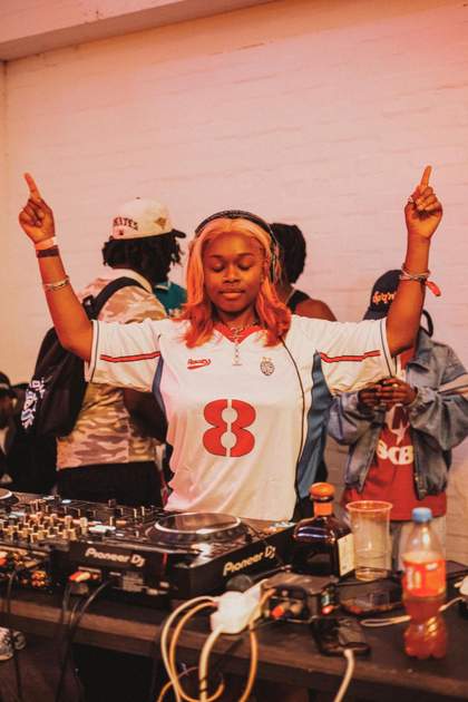 Person wearing a football shirt and headphones, standing in front of a DJ deck with their arms up