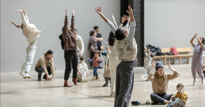 A group of people in a large grey room, some sitting on the floor and some standing with their arms outstretched to the sky