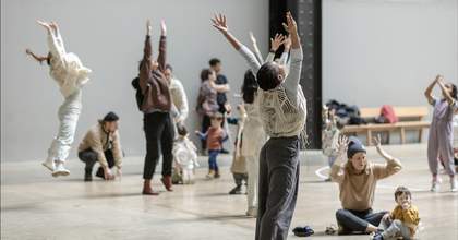 A group of people in a large grey room, some sitting on the floor and some standing with their arms outstretched to the sky