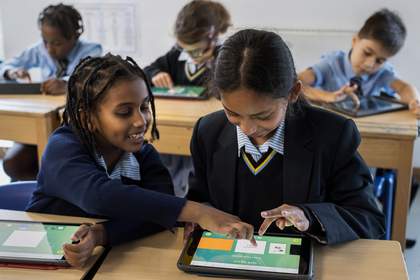 Two students sat at desks in a classroom are exploring a drawing game on tablet devices.