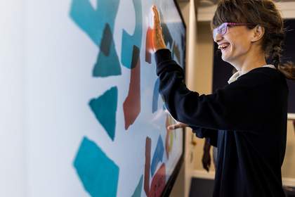 A student is standing and interacting with large coloured shapes on a large interactive whiteboard in a classroom.