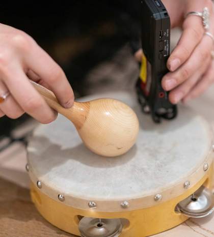 A teacher taps a tambourine with a maraca and records the sound into a portable recording device.
