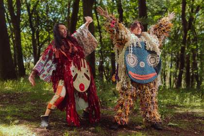 Image shows artists Frances Daykin and Huhtamaki Wab dressed in fantastical costumes they have created from Scrapstore materials. They are striking poses with arms outstretched in a sunny outdoor setting, surrounded by trees.