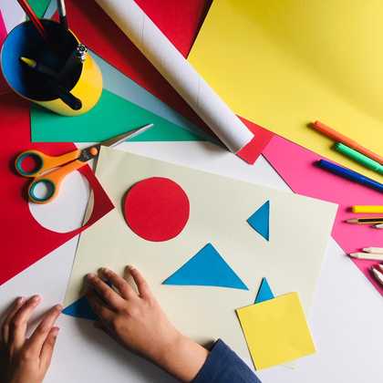 a pair of children's hands moving collage shapes around, red circle blue triangles and yellow square