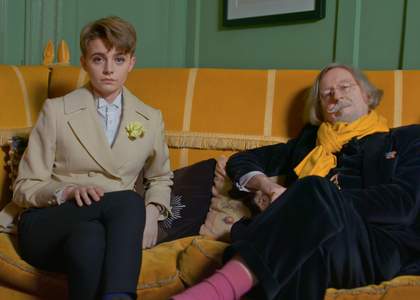 two figures in elegant dress sit on a yellow sofa