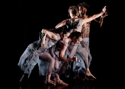 4 dancers are on an all-black stage. They wear loose-fitting sheer grey clothes. The 2 female dancers in front are side-on and crouched down, bending at the knee and upper bodies tilted forward. The front-most dancer holds 2 crutches.
