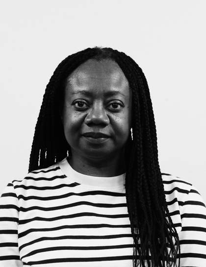 A portrait of Dianne Minnicucci in black and white. She faces the camera and has a slight smile. Her hair is in braids and parted in the centre. She is wearing a white top with black horizontal stripes.